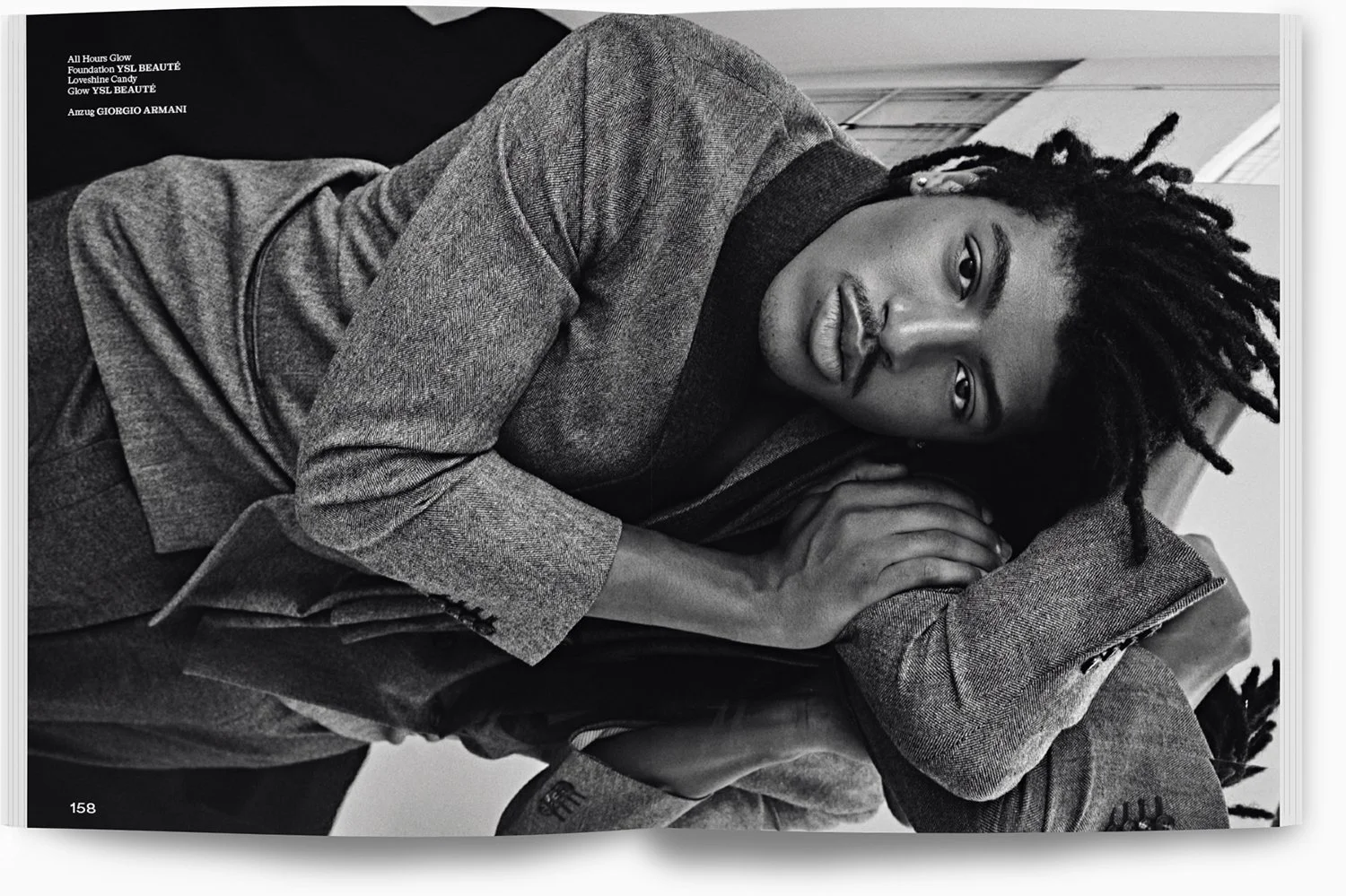 schwarze und weiße Fotografie eines Mannes mit Dreadlocks. Er liegt auf einem Arm, trägt einen Anzug aus Wolle. Blick in die Kamera, nachdenklich. Hintergrund unscharf.