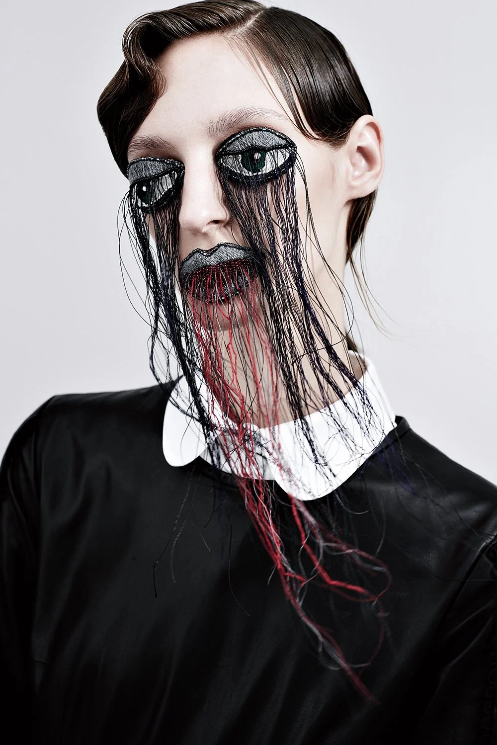 Kunstvolles Porträt einer Frau mit makeup-inspiriertem Maske, die schwarzen, grauen und roten Fäden und Stickereien zeigt, die wie fließende Tränen und Lippen aussehen.