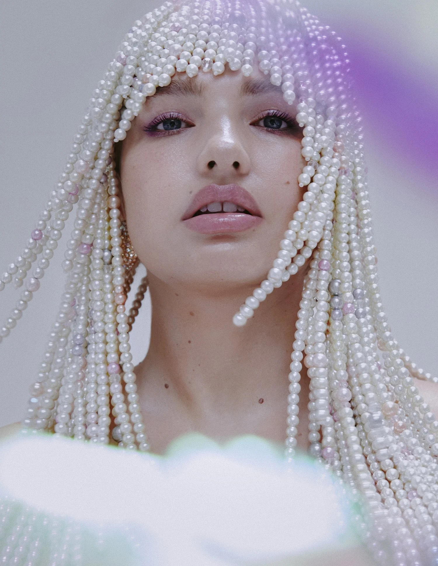 A woman with long pearl-studded braids wearing a pearl cap, with light makeup and slightly parted lips, looking directly at the camera.