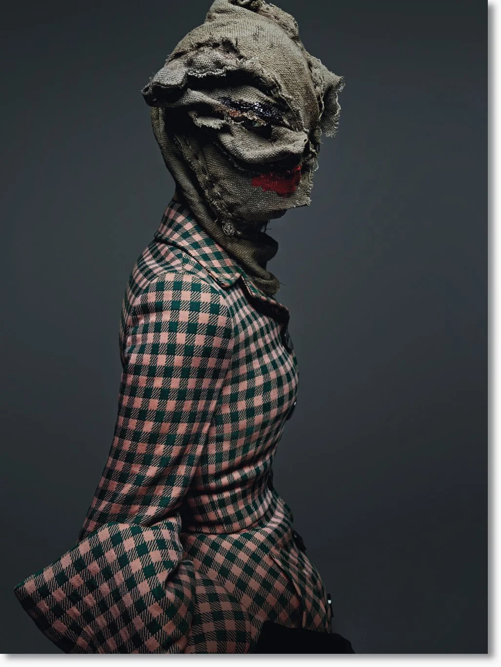 Person mit verwüstetem Stoffmaskengesicht, trägt karierten Kleidung, vor dunklem Hintergrund.