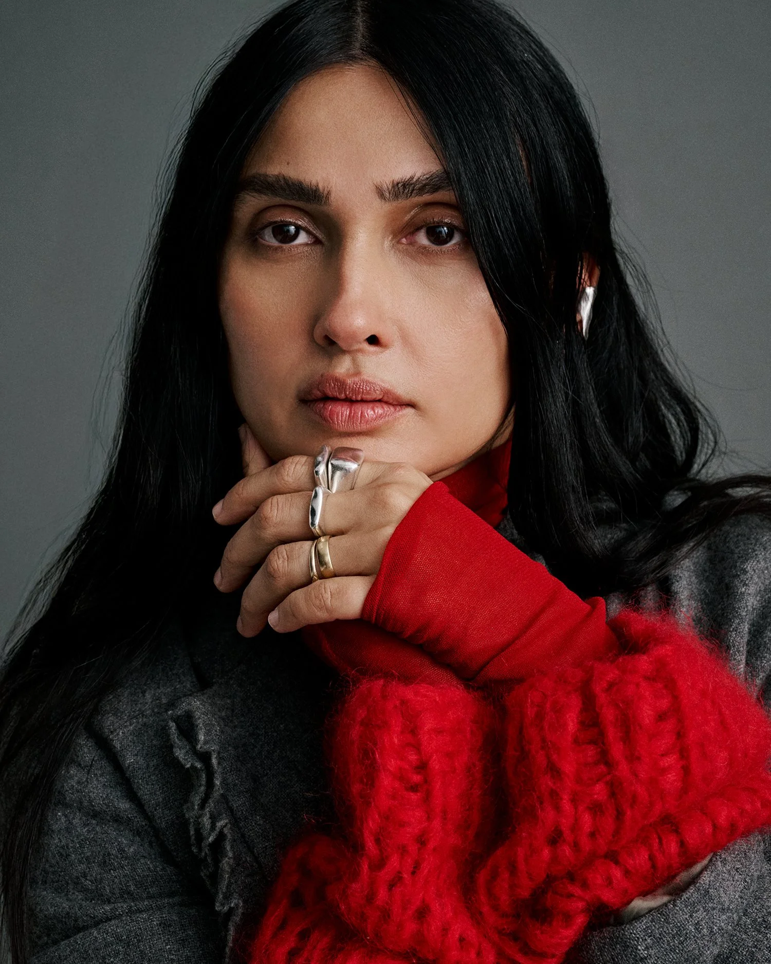 A woman with black hair and light makeup wearing a gray jacket, red sweater, and large silver rings, posing with her hand near her chin against a gray background.