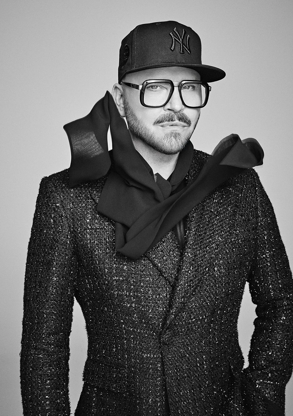 Schwarz-Weiß-Foto eines Mannes mit Glatze, Bart und Schnurrbart, der eine New Yorker Baseballcap, große Brillen, einen dunklen Schal, einen glitzernden schwarzen Mantel und schwarze Handschuhe trägt.