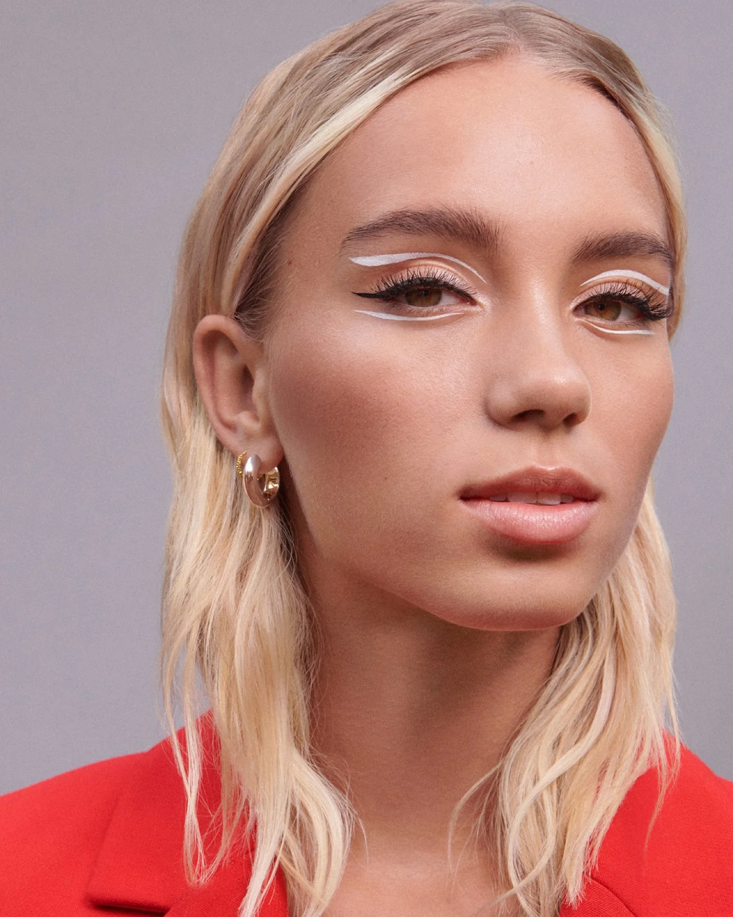 A young woman with blonde hair styled in loose waves, wearing gold hoop earrings and a red blazer. She has white eyeliner accents on her eyes and pinkish lips, with a neutral grey background.