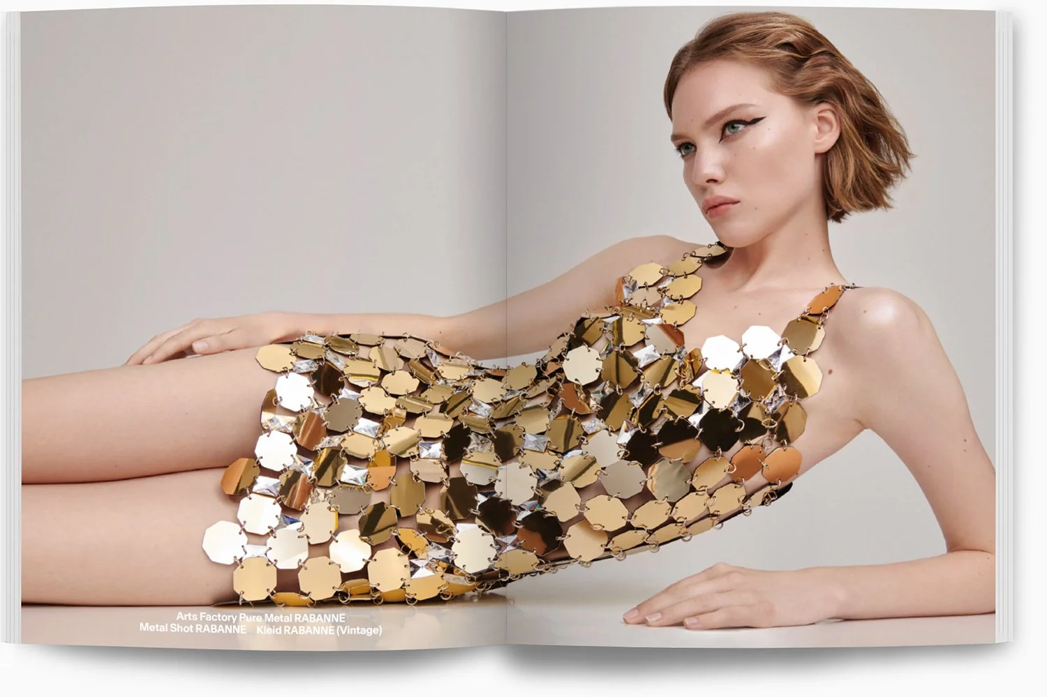 Frau in einem Kleid aus großen goldenen und silbernen Metallschalen, die wie ein futuristisches Kleid aussehen, während sie liegend auf einem weißen Hintergrund posiert.