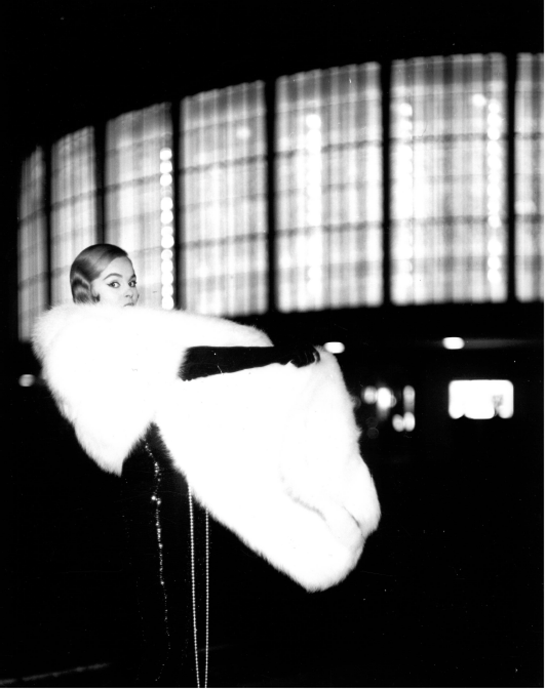Eine elegante Frau in einem langen schwarzen Kleid mit einem weißen Pelzmantel sitzt in einem Raum mit großen fensterartigen Strukturen im Hintergrund.