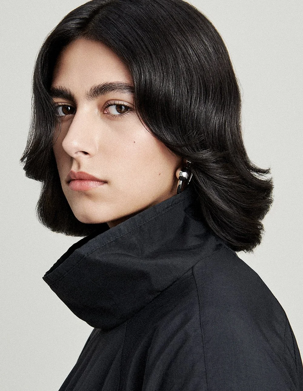 Nahaufnahme eines Mädchens mit schwarzen, mittellangen, gewellten Haaren, schwarzen Kleid mit hohem Kragen und silberner Ohrring.