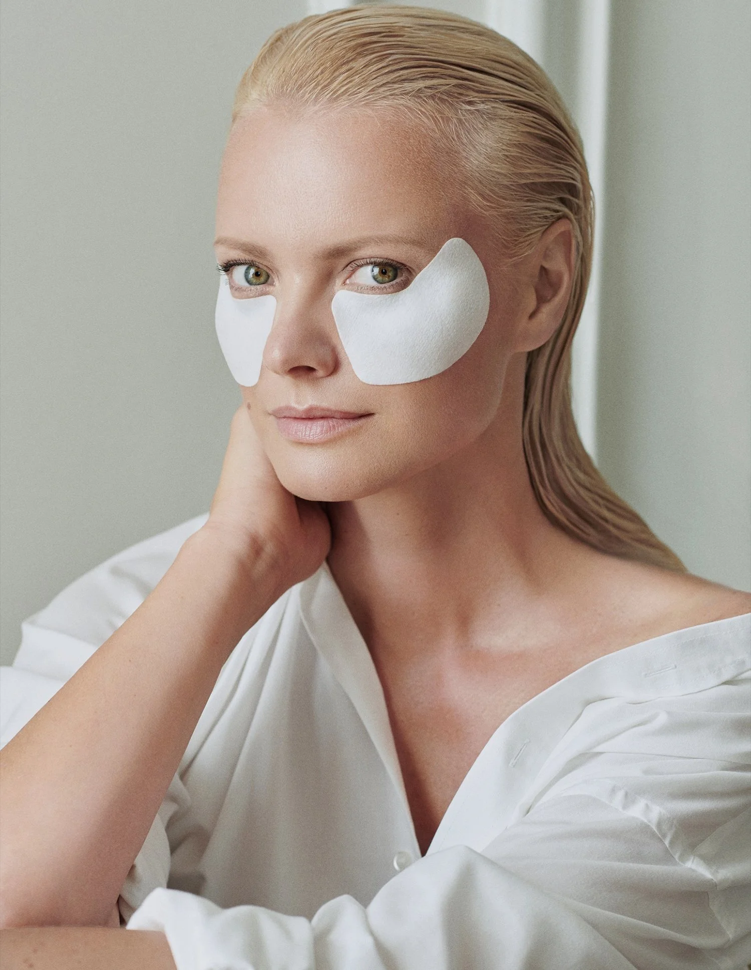 A woman with blonde hair and green eyes is wearing white eye patches under her eyes, with her hand resting on her neck, and she is dressed in a white top.