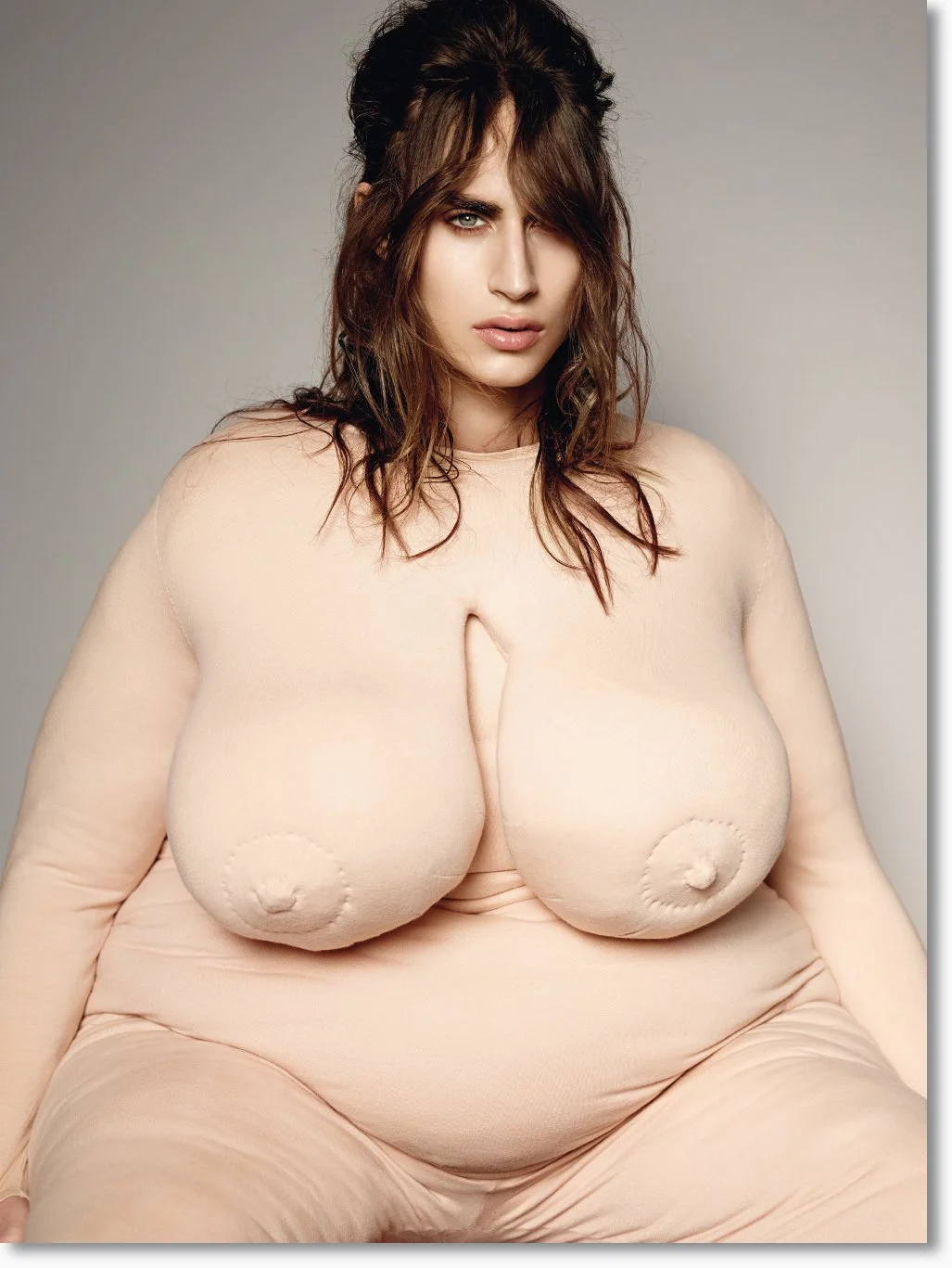 Frau mit braunen, gelockten Haaren in einem nahtlosen, hautfarbenen Kleid mit prominenten Oberarmen und vollem Dekolleté.