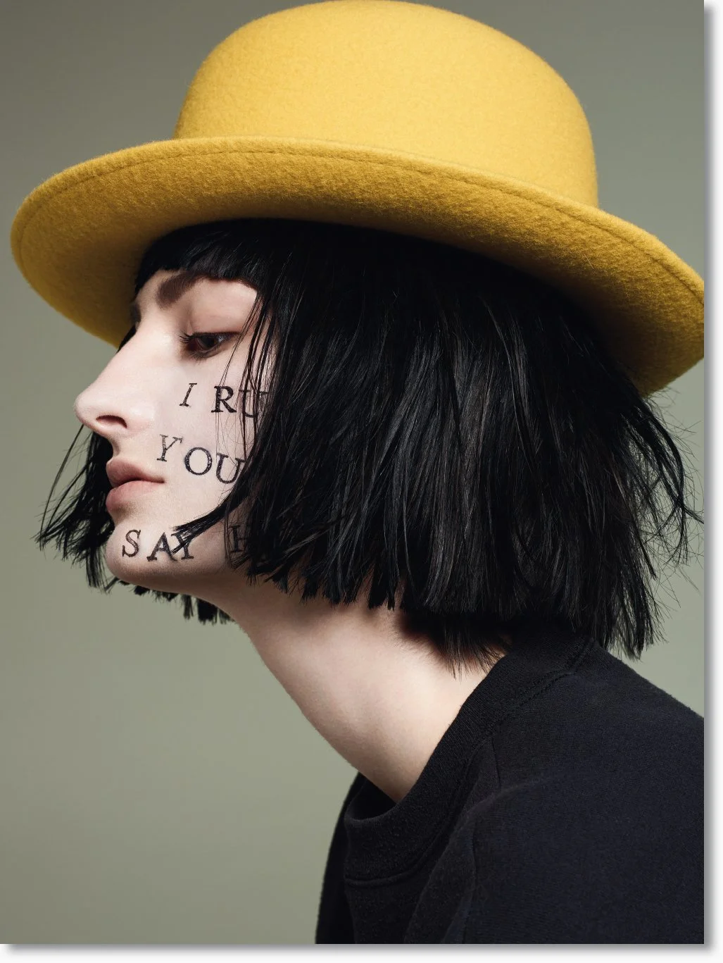 Seitenansicht einer jungen Frau mit schwarzer, kurzgestylter Frisur, trägender gelber Hut und einem schwarzen Oberteil. Auf ihrem Gesicht sind schwarze Buchstaben geschrieben.