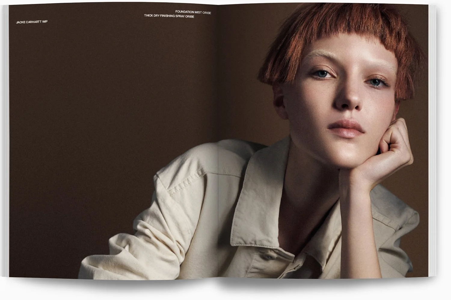 Nahaufnahme eines jungen Mädchens mit roten Haaren und blauer Augen, getragen in einem beige Hemd, lehnt mit ihrer Hand an ihrem Kiefer, vor einem dunklen, einfarbigen Hintergrund.