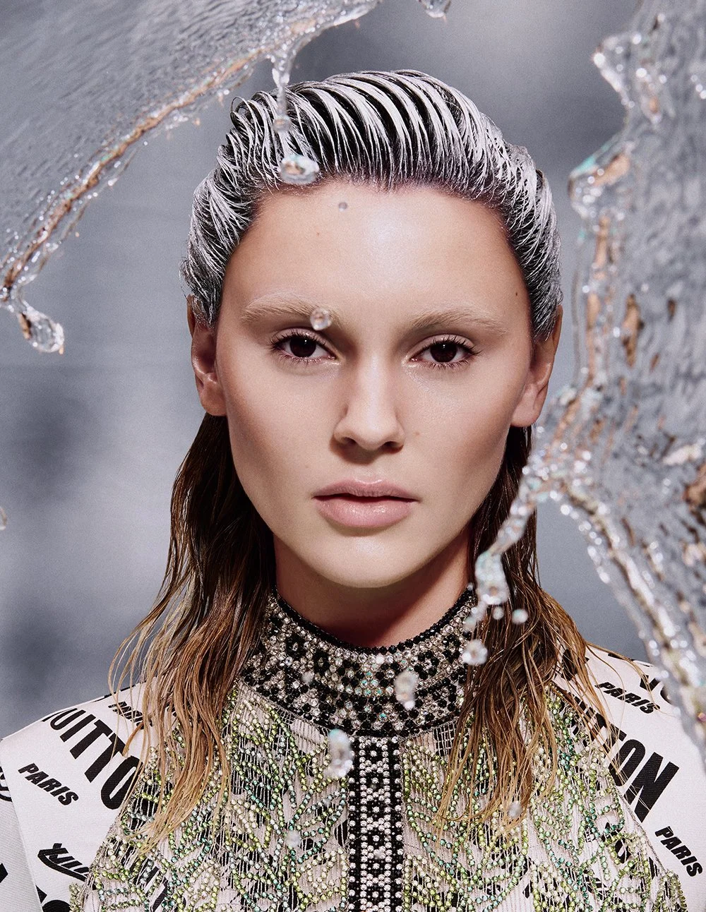 Eine Frau mit nassen, zerzausten Haaren, die mit Wasser bedeckt sind, trägt ein schimmerndes, visuell auffälliges Outfit mit Pearls und Glitzer, und ist vor einem grauen Hintergrund. Sie blickt direkt in die Kamera.