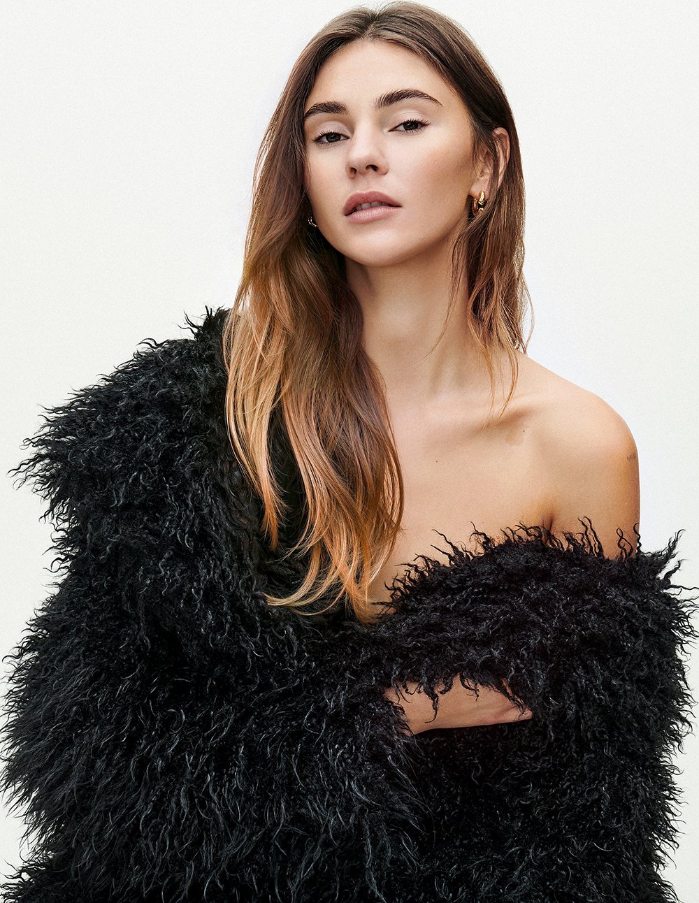 Ein junges Mädchen mit langem, braunem Haar, das teilweise gewellt ist, trägt eine schwarze, pelzige Jacke und schaut in die Kamera. Sie hat goldene Ohrringe und steht vor einem weißen Hintergrund.