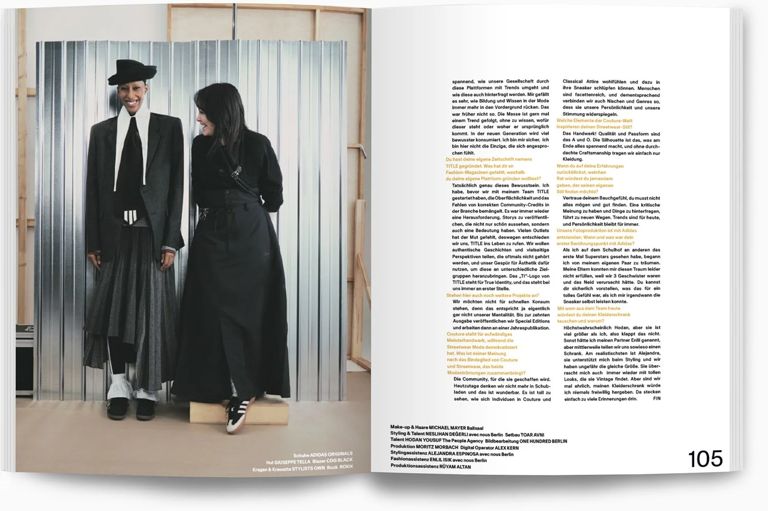 Zwei Frauen in Business- oder Modekleidung, die in einem Fotostudio vor einer metalldornen Wand stehen und lachen, während sie für ein Mode-Fotoshooting posieren.