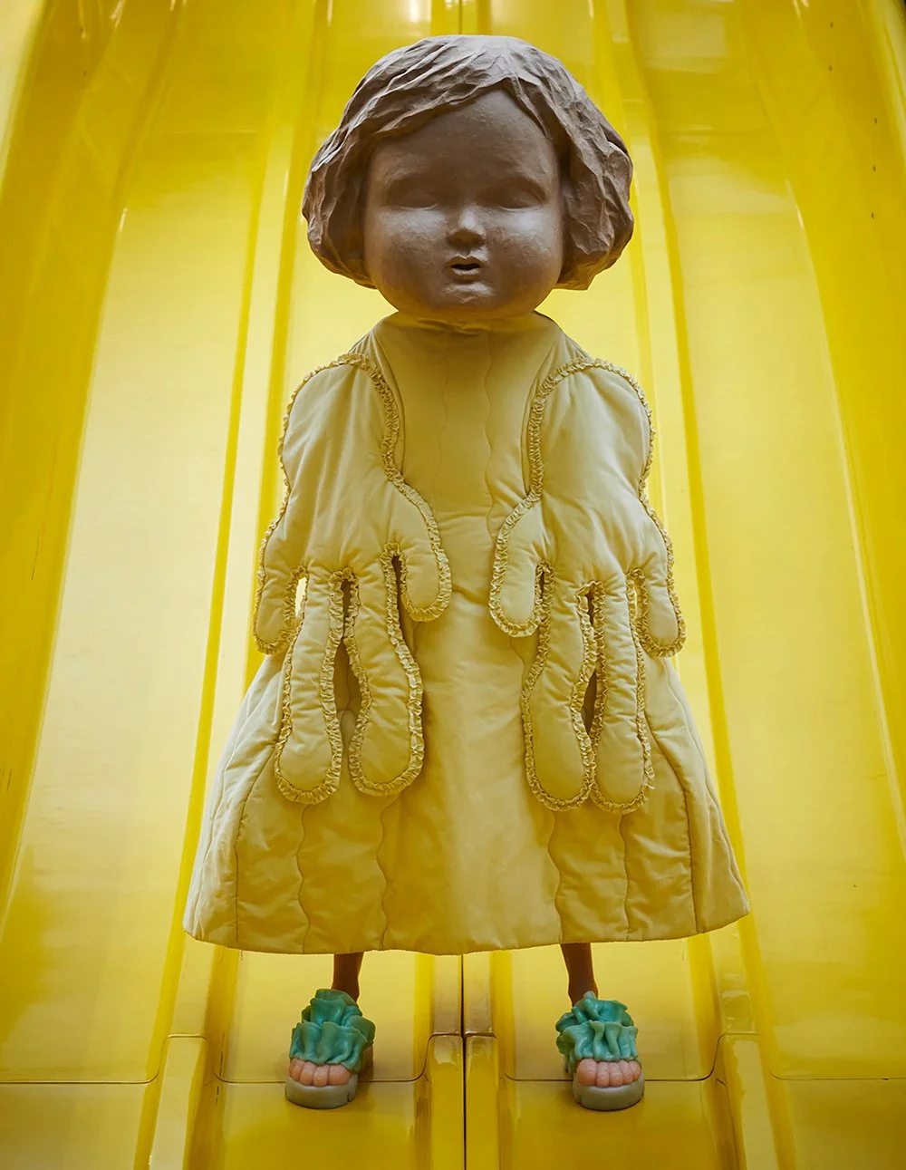 A large art installation of a girl with a doll-like face, wearing a yellow dress with multiple tentacle-like appendages, on a yellow slide.