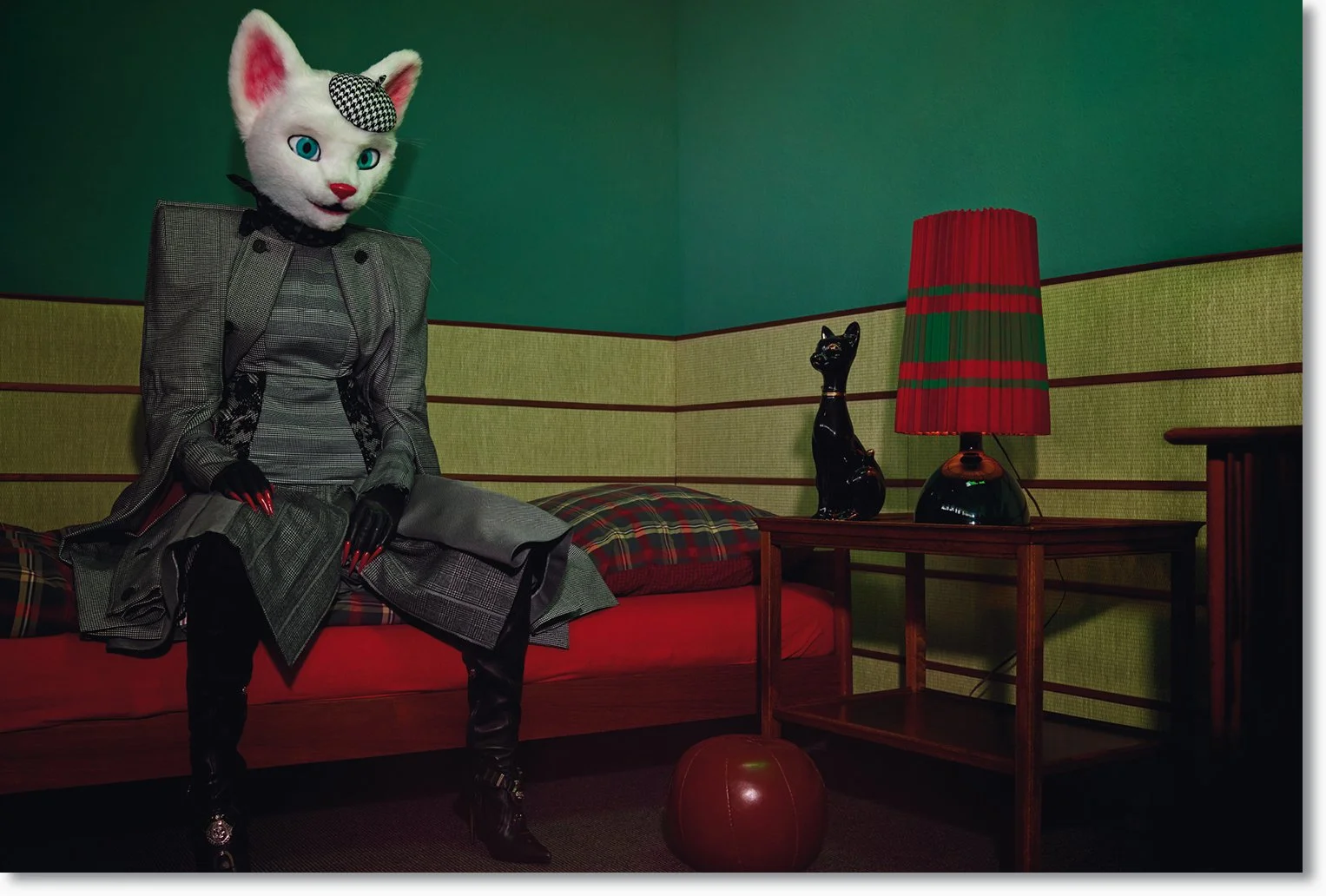 Eine völlig menschlich gekleidete Person mit einer Katzenmaske sitzt auf einem roten Sofa in einem Raum, der mit grüner Wand und decorativen Elementen eingerichtet ist.