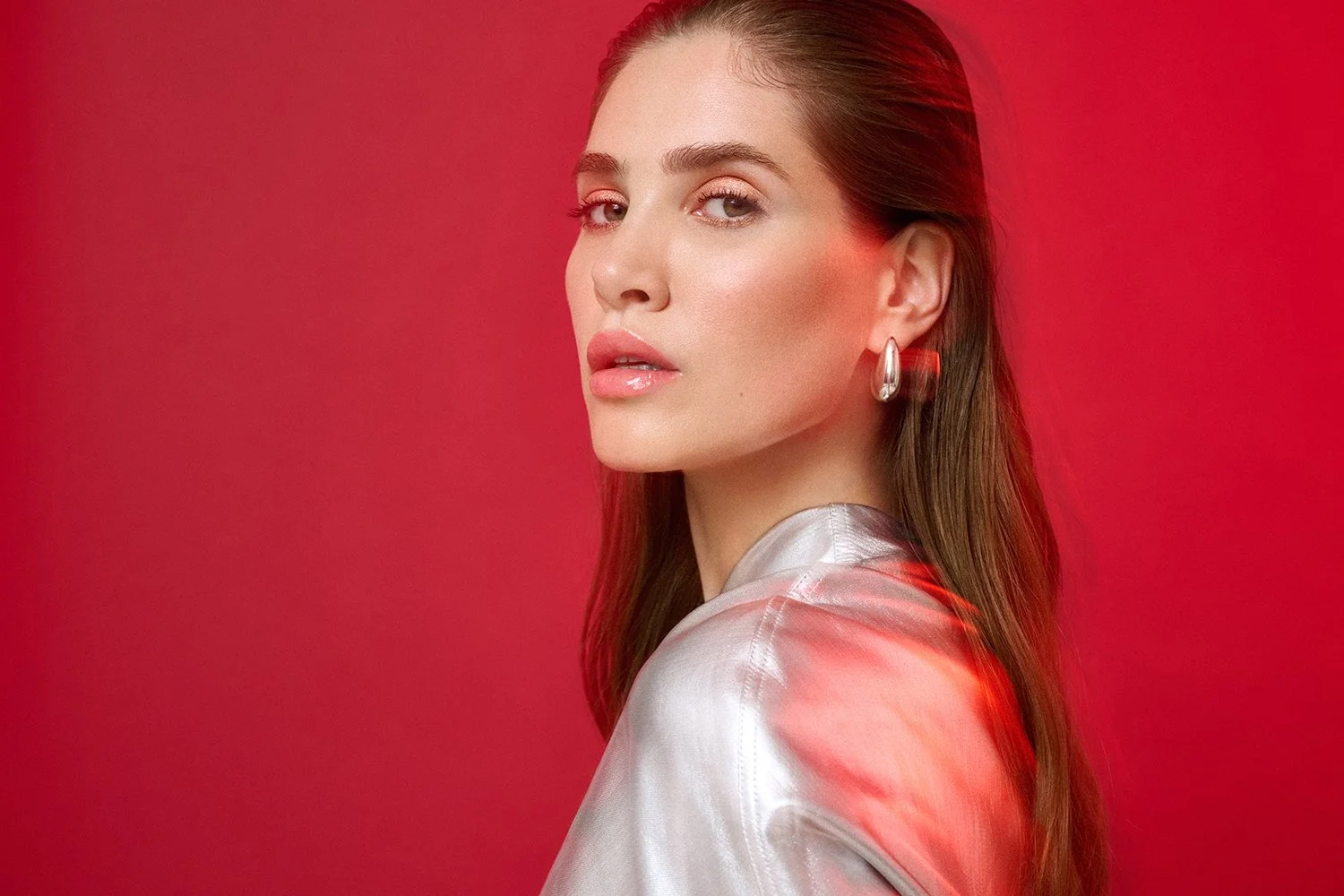 A woman with long brown hair wearing a shiny silver top and silver hoop earrings, looking over her shoulder against a red background.