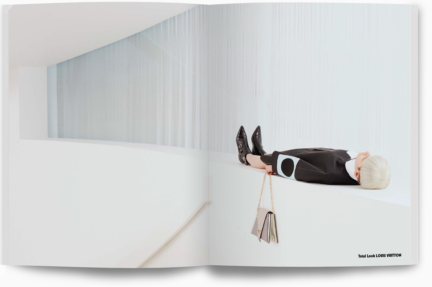 Eine Person mit blonden Haaren liegt auf einem weißen Sims vor einer weißen Wand in einem minimalistischen Raum, trägt schwarze Schuhe, schwarzen Rock, weißen und schwarzen Oberteil, hält eine Tasche mit Goldkette und hängt an der Wand eine beige Tas