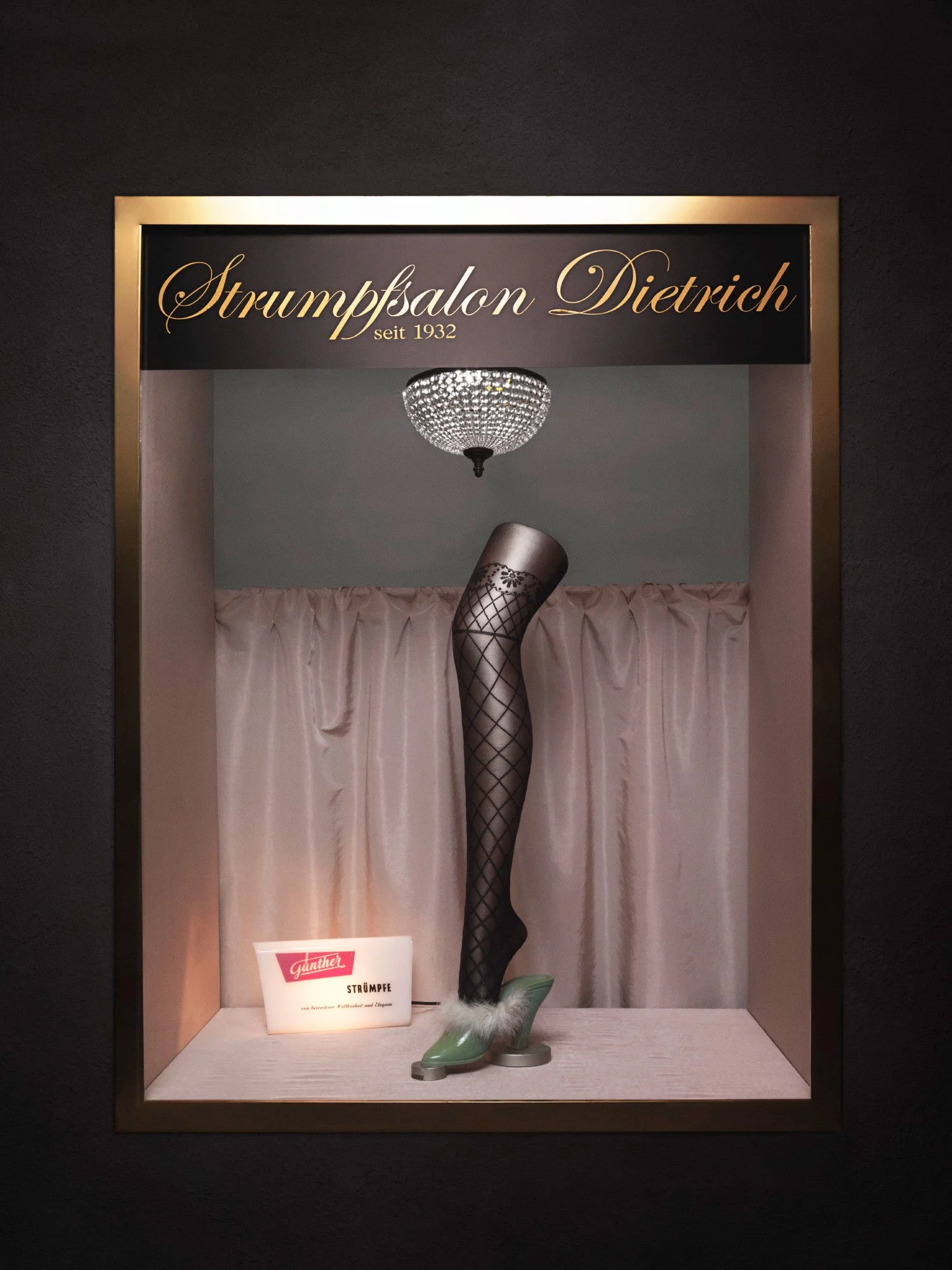 A display case showing a mannequin leg in fishnet stockings and green high-heeled shoes, with a chandelier overhead and a pink curtain background, with a sign featuring the Günther logo and the word 'Strümpfe'.