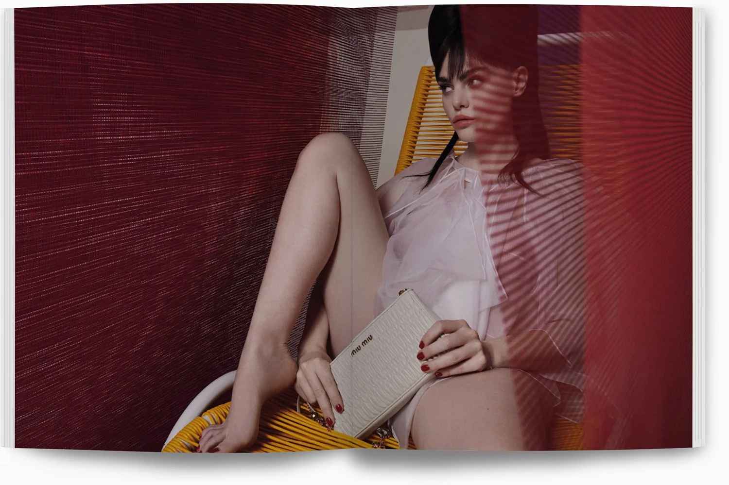 Frau sitzt auf einem gelben Stuhl, hält eine weiße Miu Miu Geldbörse, trägt ein transparentes weißes Oberteil, hat dunkle Haare und rote Nägel, blickt nach rechts, im Hintergrund rot-gestreifte Wand. 