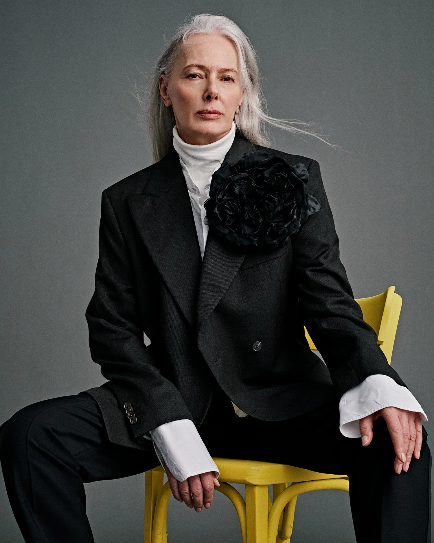 A mature woman with long gray hair sitting on a yellow chair against a gray backdrop, dressed in a black suit with a large black fabric flower on her shoulder, white shirt, and turtleneck, with a serious expression.