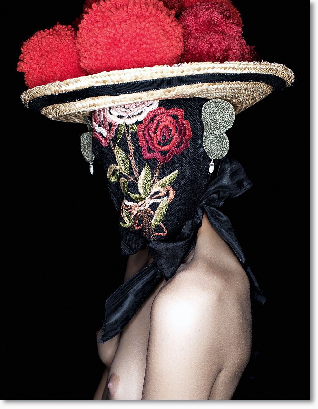 Person trägt großen Hut mit roten Pompons, schwarze Bandana mit Rosenstickerei, große Ohrringe, obere Körperhälfte ohne Kleidung.