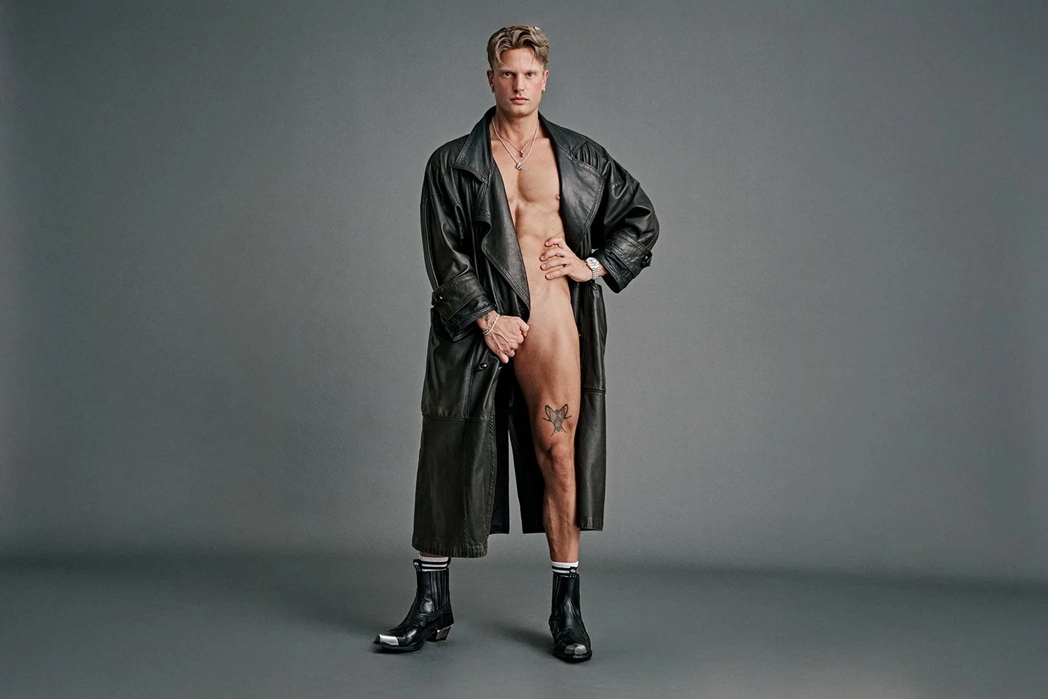 A young man standing against a plain grey background, wearing a long black leather coat, black heeled boots, and striped socks. He is topless, with one hand on his hip and the other holding the coat open, revealing a tattoo on his thigh.