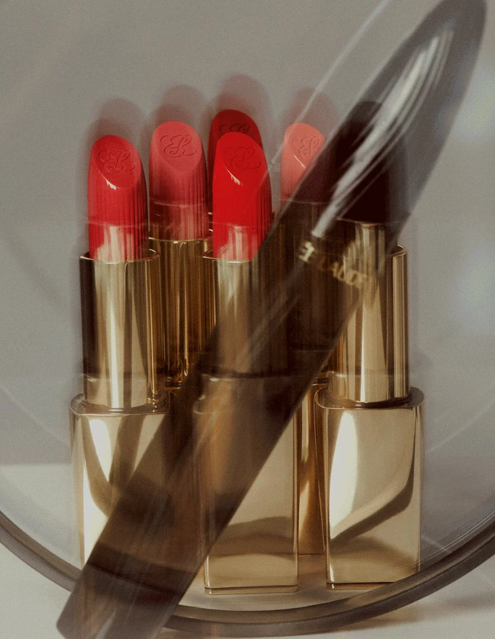 Red lipsticks in a gold lipstick holder, with a partially visible black lipstick tube leaning on them, inside a glass container.
