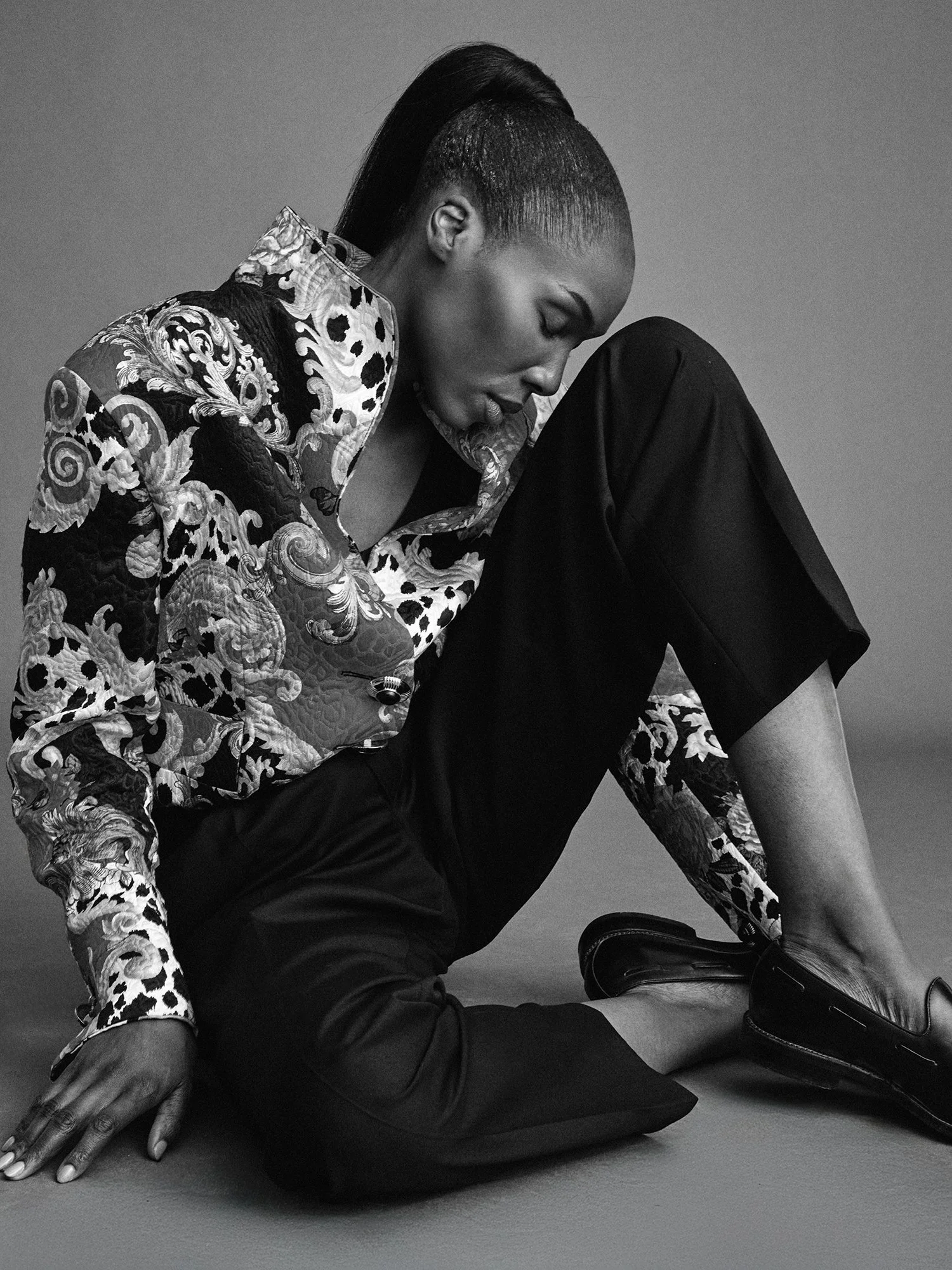 Black and white photo of a woman with short hair in an intricate patterned blouse and black pants, sitting on the floor with one leg bent and foot near her head, looking down with eyes closed.