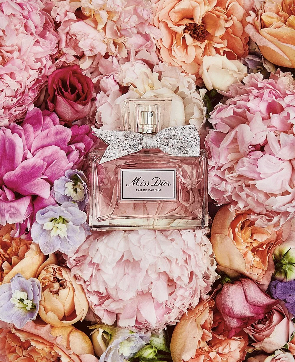 A bottle of Miss Dior Eau de Parfum surrounded by pink, peach, and lavender flowers.