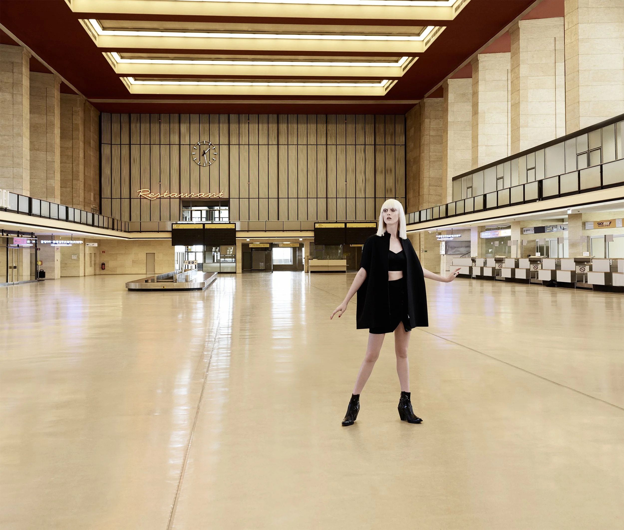 Eine junge Frau mit weißen Haaren trägt einen schwarzen Mantel, schwarzen Rock und schwarze Stiefel, steht in einem großen, leeren, lichtdurchfluteten Bahnhof oder Flughafen mit offenem Raum, Holz- und Steinverkleidungen, einer Uhr an der Wand und ei