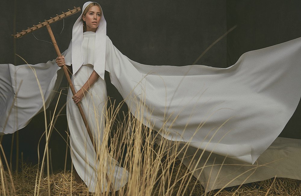 Eine Frau im weißen Gewand hält eine Hacke, umgeben von hohen Gräsern, vor einem dunklen Hintergrund.