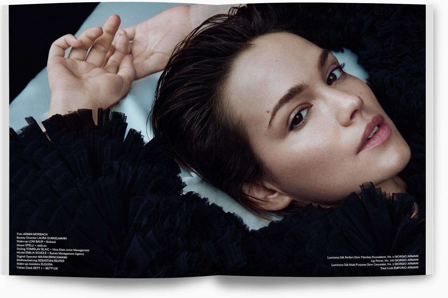 Nahaufnahme eines liegenden Models mit dunklem, glattem Haar, dezentem Make-up, schwarzen Textilkleid mit Rüschen und natürlichem Ausdruck, im Hintergrund dunkel und hellblaues Kissen.