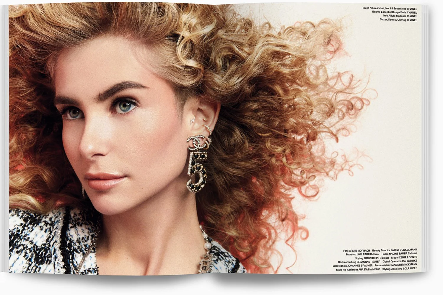 Eine Frau mit lockigem, rotem Haar trägt auffällige Ohrringe und ein Mode-Outfit, Blick leicht zur Seite, vor hellem Hintergrund.