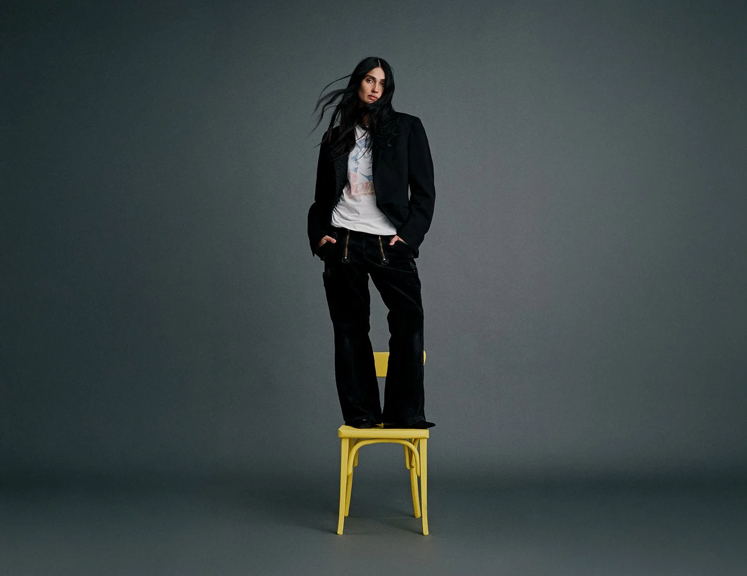 A woman in black pants, a black blazer, and a white T-shirt standing on a yellow chair against a gray background.