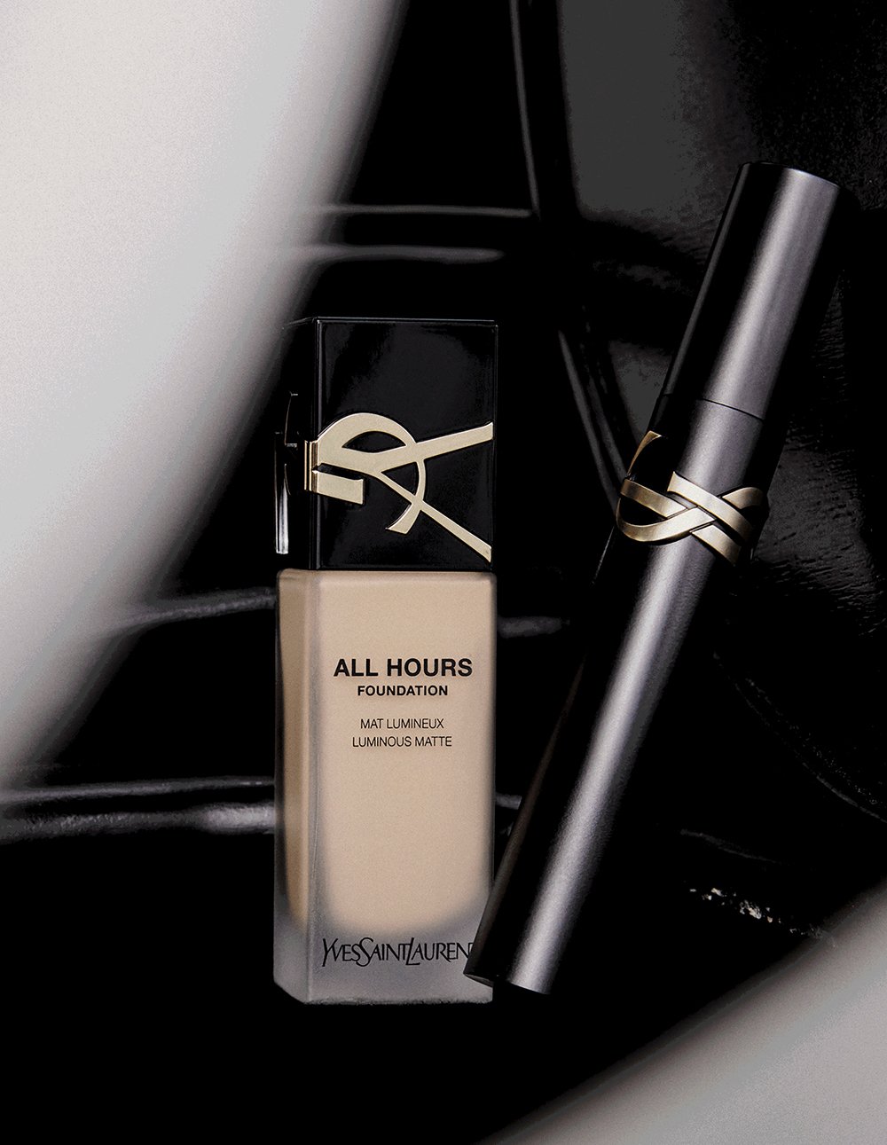 A bottle of Yves Saint Laurent All Hours Foundation with a black cap and beige liquid, a sleek silver tube with a black cap, and a black Yves Saint Laurent box, all set against a dark background.