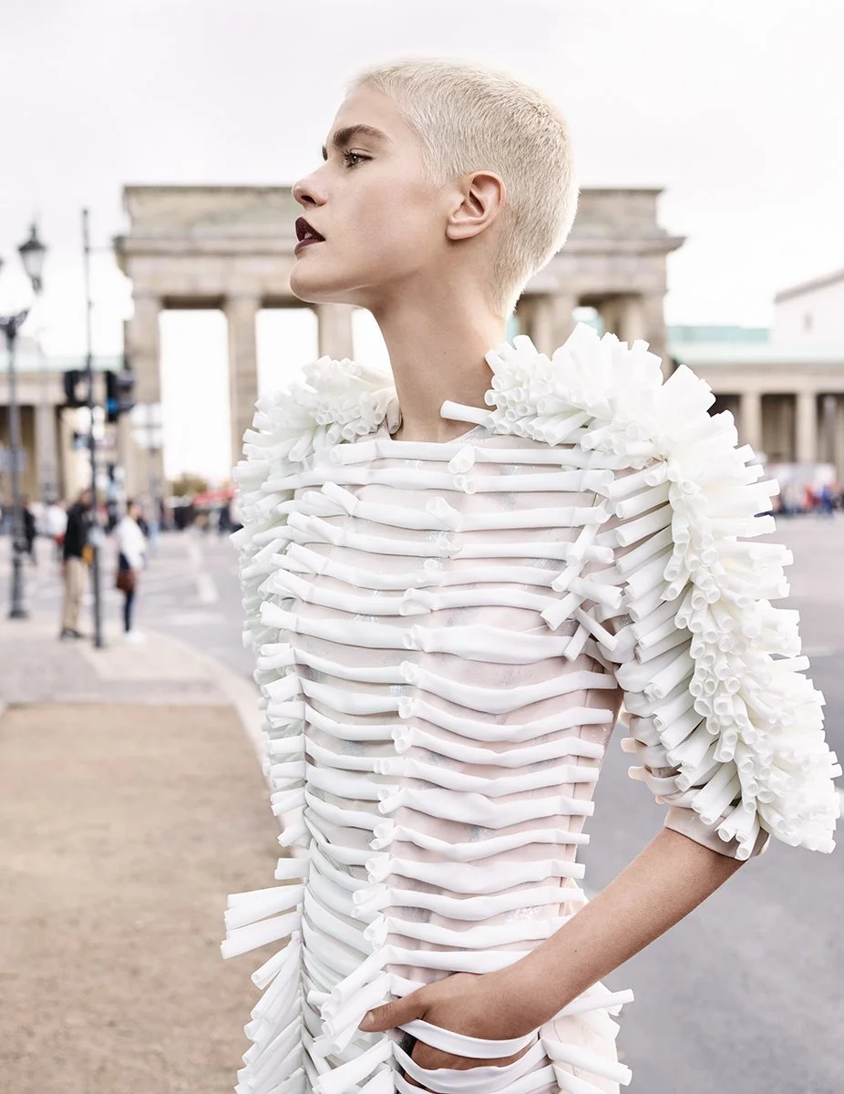 Nahaufnahme einer Frau mit kurzem blonder Haar, die ein futuristisches, weißes Kleid mit einer Struktur aus Röhren trägt. Im Hintergrund ist das Brandenburger Tor in Berlin zu sehen.