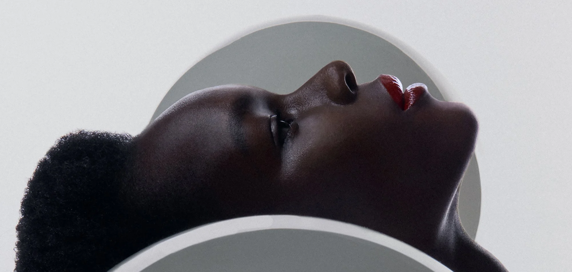 Porträt einer schwarzen Frau mit geschlossenen Augen, roten Lippen und auf dem Kopf einem schwarzen Lockenkopf, liegend in einer weißen runden Architektur.