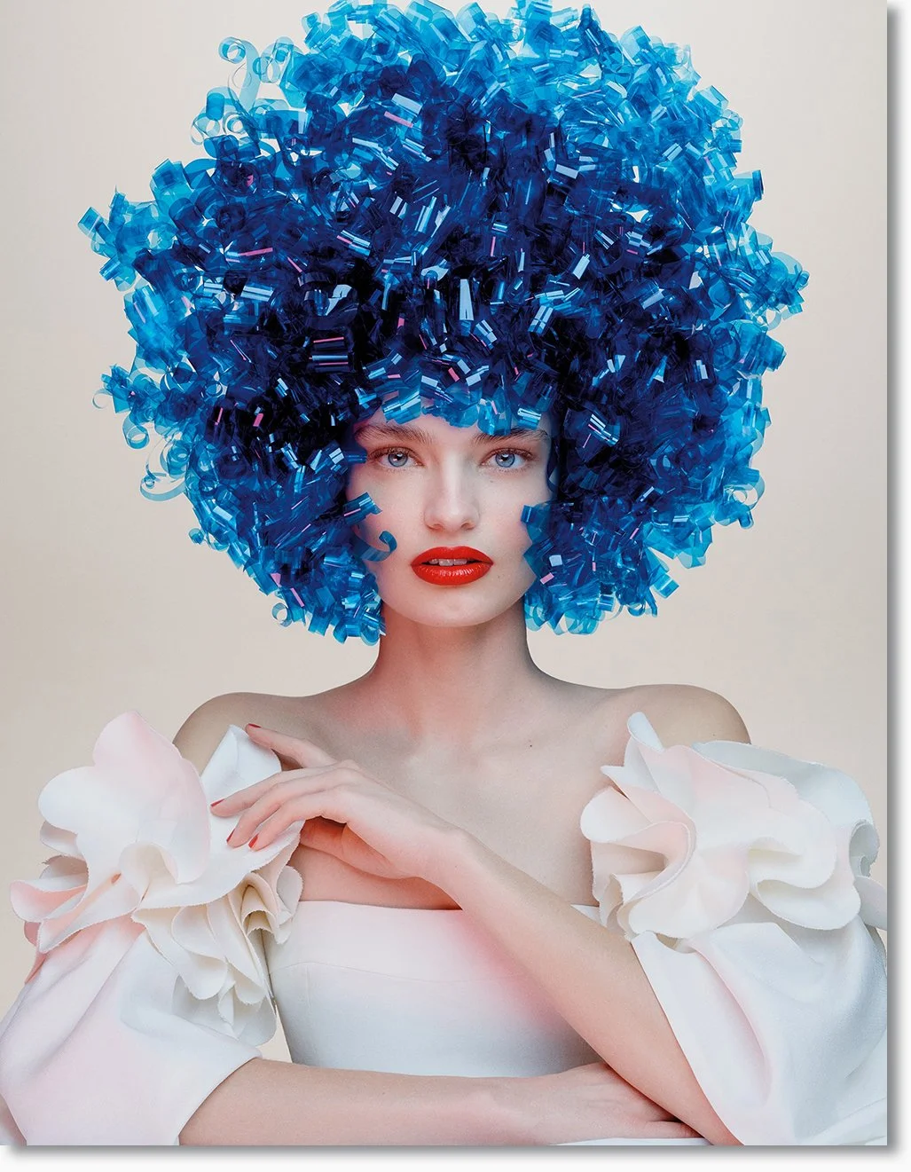 Eine Frau mit einem großen, blauen, lockigen Hut aus Konfetti und einer weißen Kleidung mit großen Blumenapplikationen.