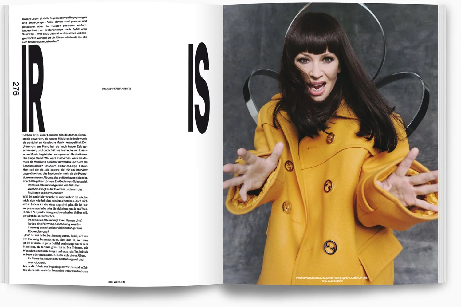 Eine Frau mit langen dunklen Haaren trägt einen goldgelben Mantel und posiert energisch für die Kamera, gestikuliert mit den Händen. Hintergrund ist grau und metallisch.