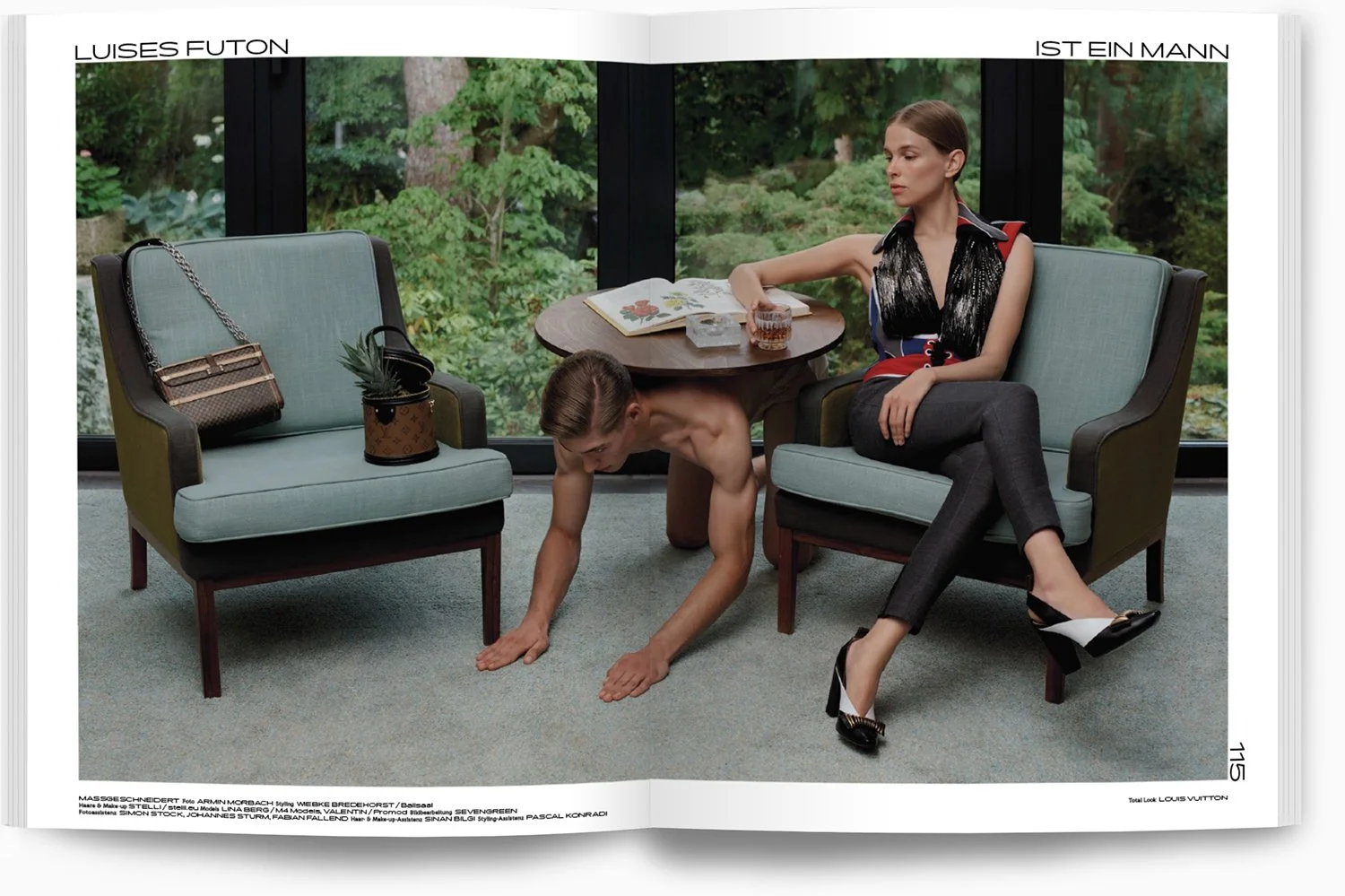 Ein Mann, der auf Händen und Füßen auf einem Teppich steht, während eine Frau auf einem Sessel sitzt, in einem Raum mit großen Fenstern und einem Garten im Hintergrund.