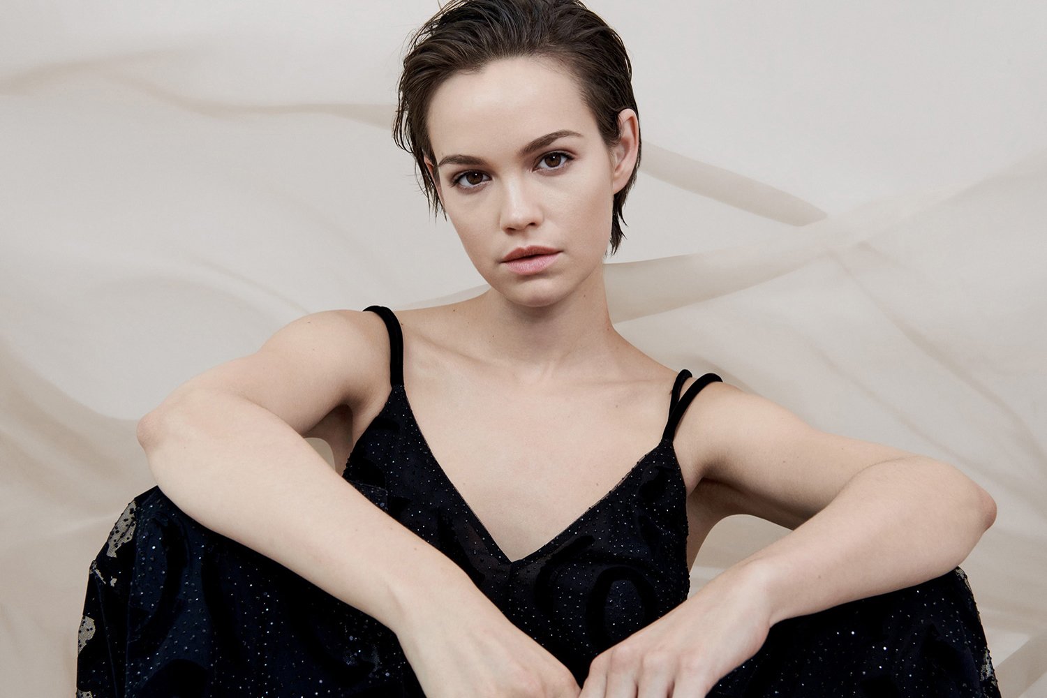 A young woman with short dark hair, wearing a black spaghetti strap dress, sitting against a light-colored abstract background.