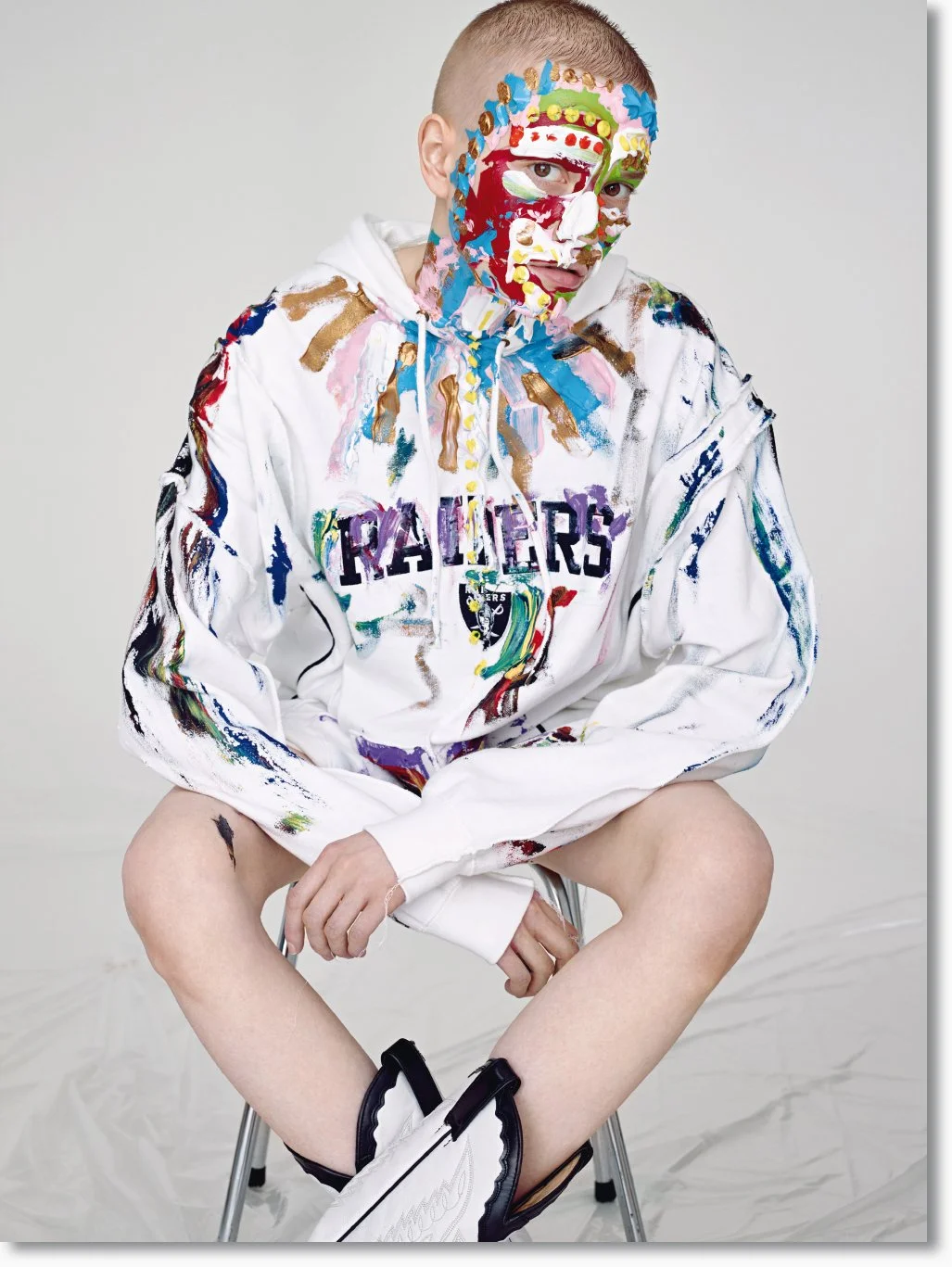 Junger Mann sitzt auf einem Stuhl, trägt ein weißes Hoodie mit gefärbten Flecken und der Aufschrift 'RAIDERS'. Sein Gesicht und Hoodie sind künstlerisch bemalt mit bunten Farben und Mustern.