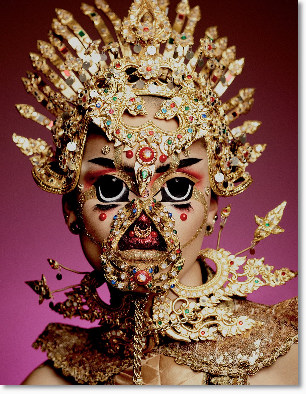 Person trägt ein aufwendig verziertes, goldenes Kostüm mit Schmuck und Kopfschmuck, das an eine hinduistische Gottheit erinnert, mit großen Augen-Make-up und traditionellen Elementen.