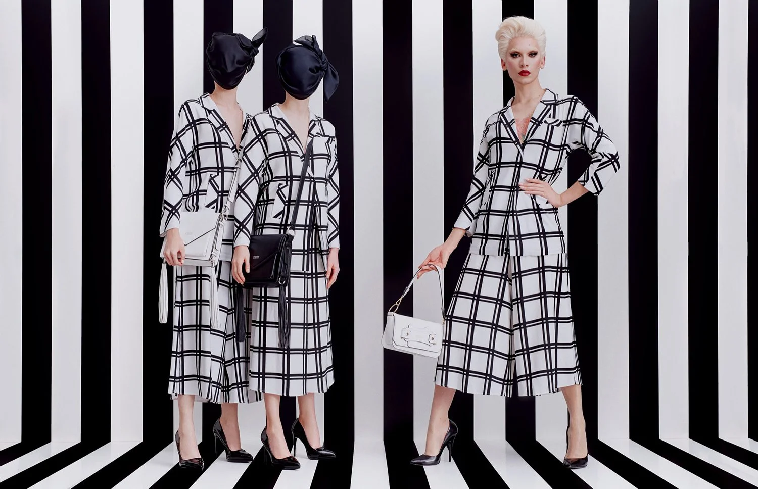 Four women in black and white plaid matching outfits, three with headscarves and bags, standing against a black and white striped background.