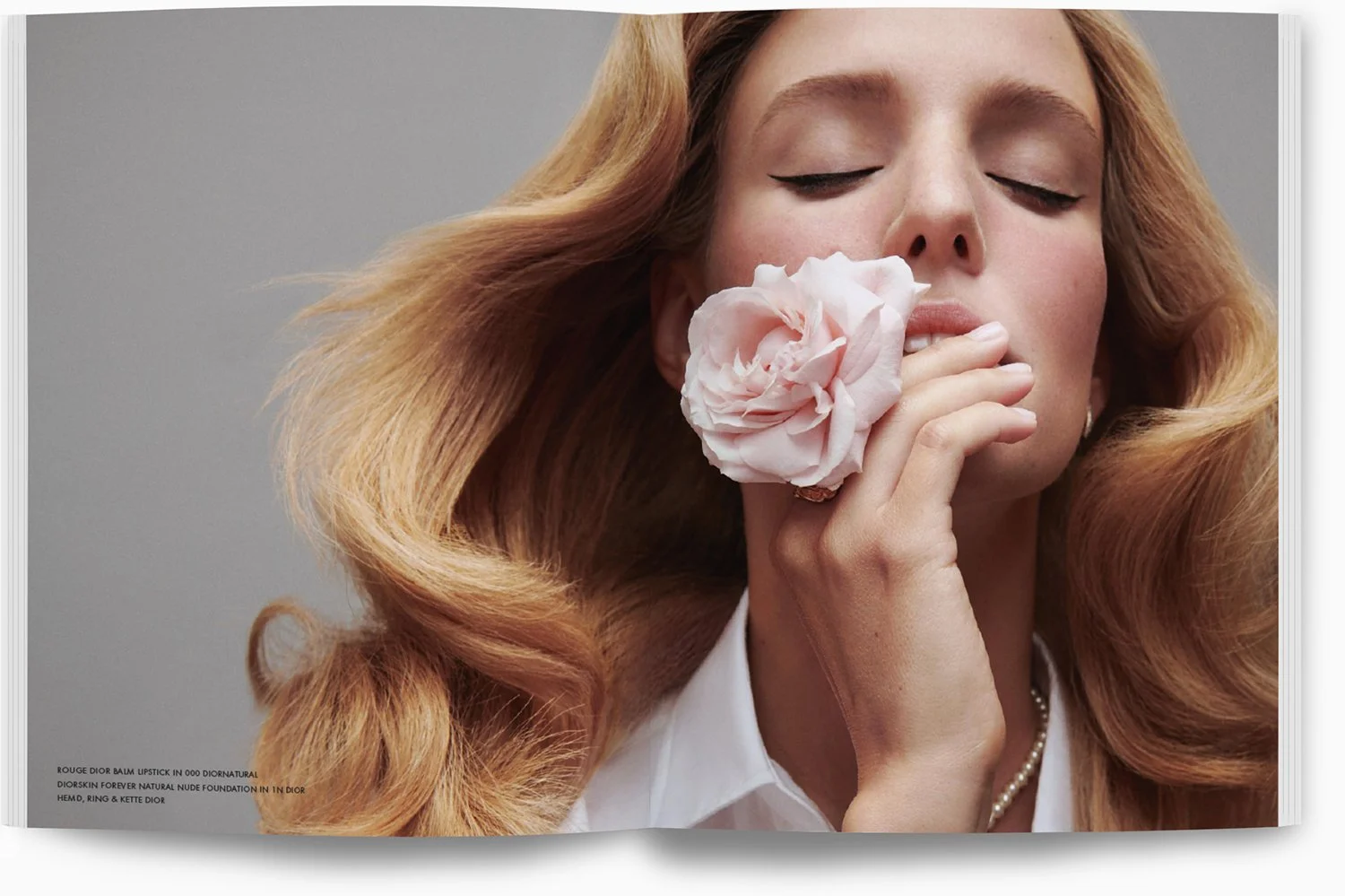 Eine Frau mit langen, lockigen, roten Haaren hält eine rosa Rose an ihr Gesicht und hat die Augen geschlossen.