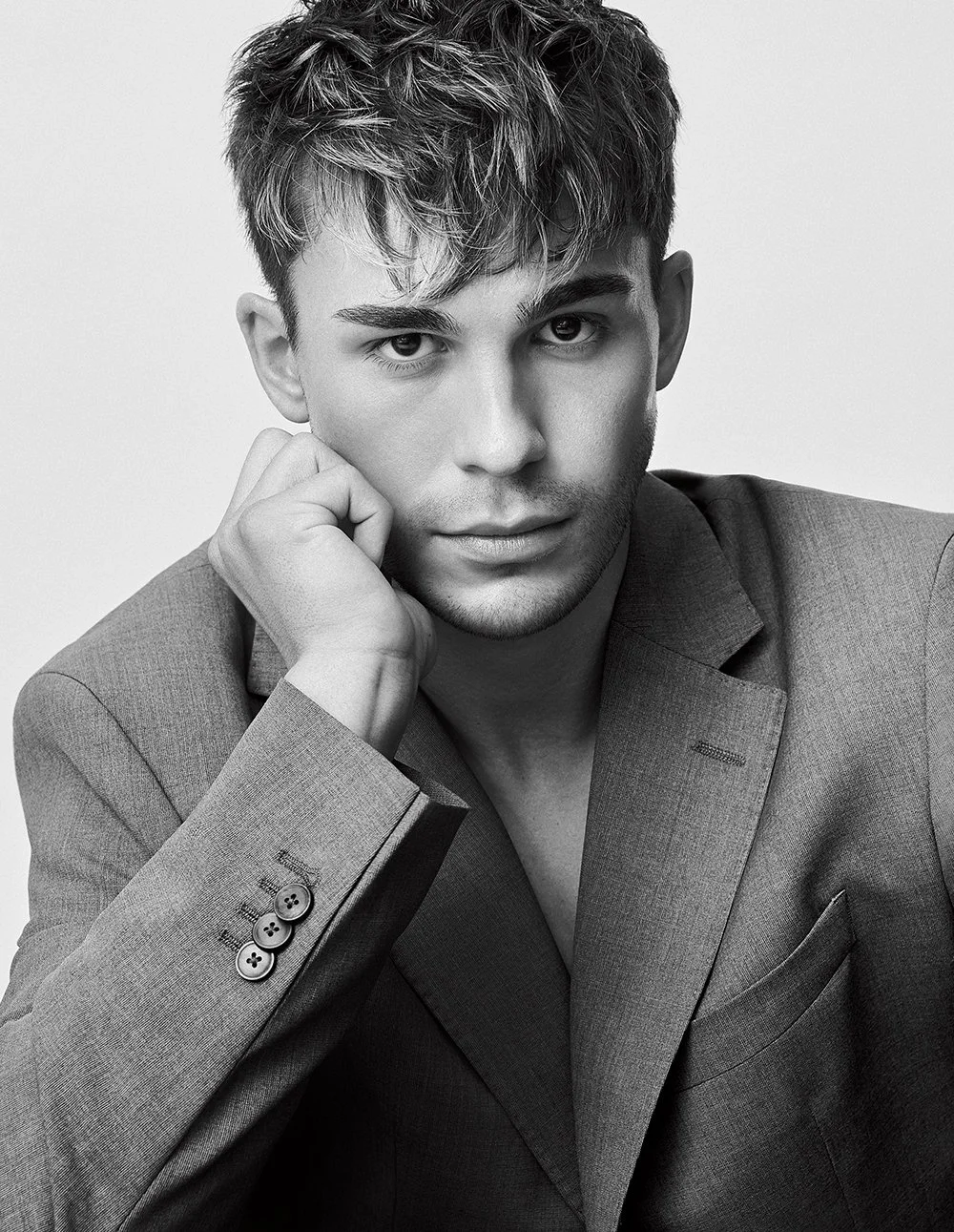 Schwarz-weiß Porträt eines jungen Mannes mit kurz gestyltem Haar, in einem Anzug, der mit der Hand sein Gesicht berührt und direkt in die Kamera schaut.