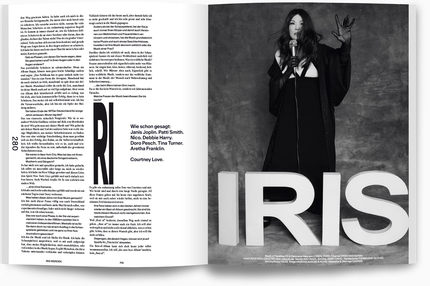 Schwarz-weiß-Foto einer Frau mit dunklem, langem Haar, die eine weiße Handkette trägt und eine Glühbirne in der linken Hand hält. Sie steht vor einer grauen Wand und trägt einen schwarzen Mantel.