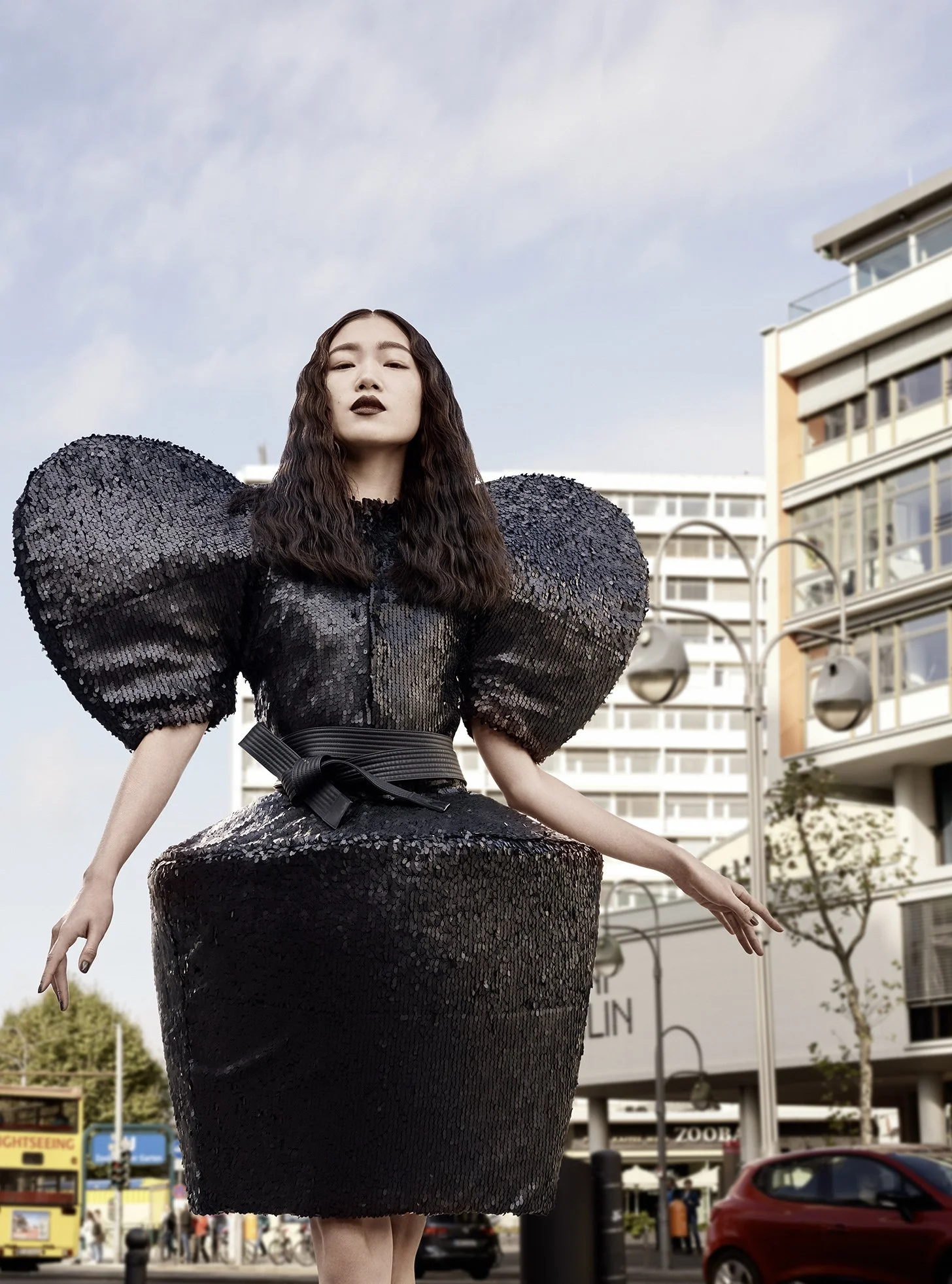 Eine Frau in einem auffälligen schwarzen Kleid mit großen, puffigen Schultern steht vor Stadtgebäuden, im Freien bei Tageslicht.