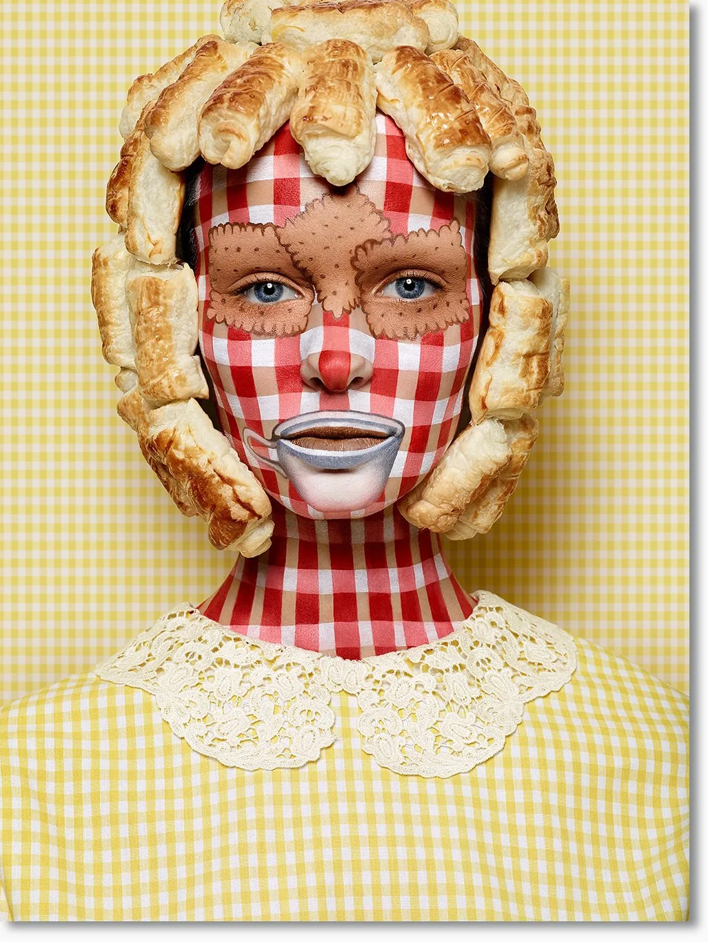 Porträt einer Person mit Bodypaint, die von Croissants umrahmt ist und einem karierten Gesichtsmuster trägt, mit großem roten Nasenstift, und einem gelben Hemd mit Spitzenkragen vor gelb-weißem Gingham-Hintergrund.