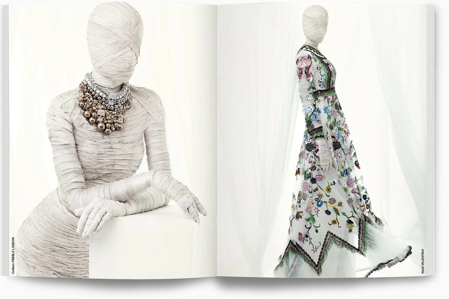 Zwei modische Kleider auf Schaufensterpuppen, eine auf der linken Seite, die mit weißen, bandartigen Stoffen umwickelt ist und große Schmuckketten trägt, die andere auf der rechten Seite mit einem langärmligen, floral bestickten Kleid vor weißen Vorh